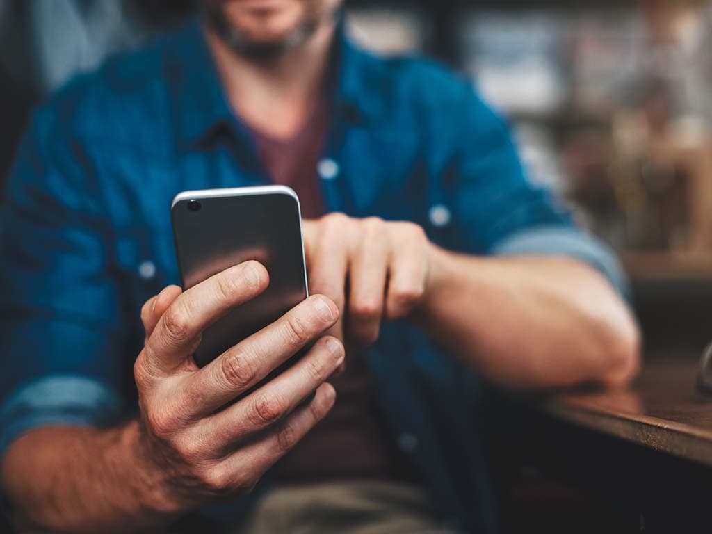 Zbliżenie mężczyzny w dżinsowej koszuli siedzącego przy stole i korzystającego ze smartfona; jego twarz jest poza kadrem, a uwaga skupiona jest na dłoniach trzymających telefon.